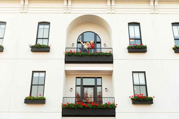 Attiucs-Hotel-Penthouse-Balcony-Exterior White multi-story building with flower boxes on each balcony; a couple stands on the upper central balcony overlooking the scene.