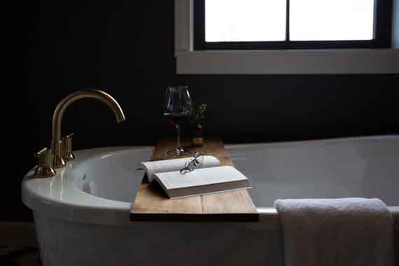 Attiucs-Hotel-Penthouse-Bathtub Open book and glasses on a wooden bath tray across a white freestanding tub, with a wine glass and towel in a dark bathroom.