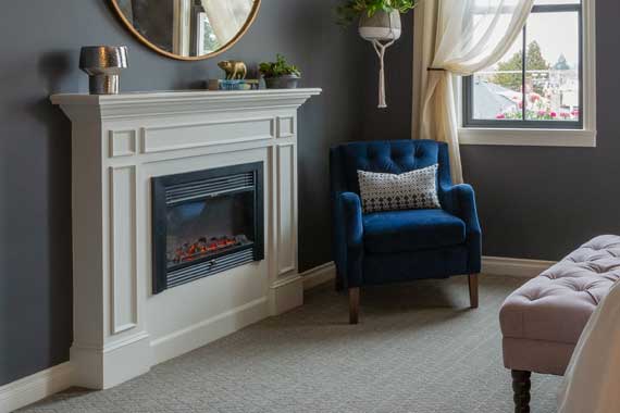 Cozy living room corner with a white mantel fireplace, a blue armchair, and a light pink tufted ottoman by a sunlit window.