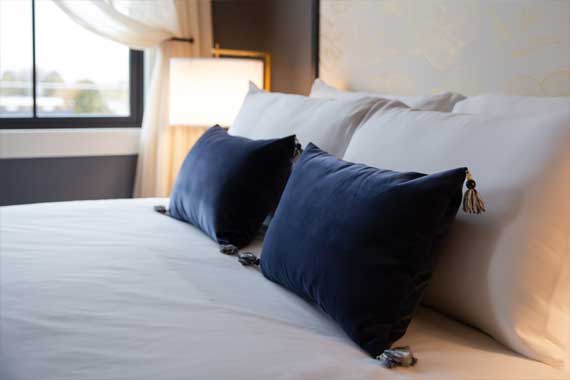 Hotel-style bed with white sheets and navy throw pillows by a window and soft bedside lamp.