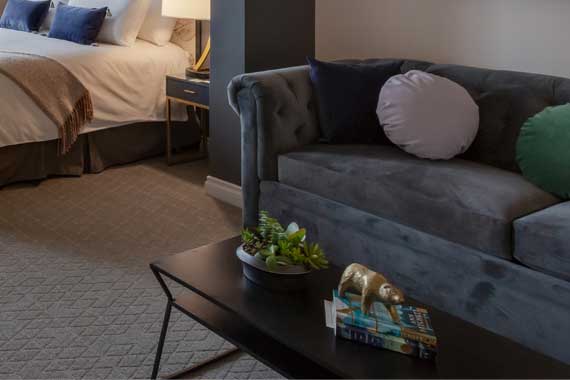 Hotel suite living area with a gray tufted sofa, round cushions, and a coffee table holding a plant and books; bed is visible in the background.