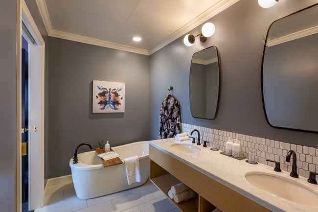 Atticus Hotel Balcony Suite Bathroom Modern bathroom with a double vanity, two oval mirrors, and a freestanding tub against gray walls; robe hangs by the sink area and artwork on the wall.