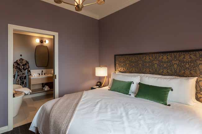 Atticus Hotel Balcony Suite Bedroom and Bathroom Hotel room with purple walls, a large bed with white linens and green pillows, and an open doorway to the en-suite bathroom showing a robe on a hook.