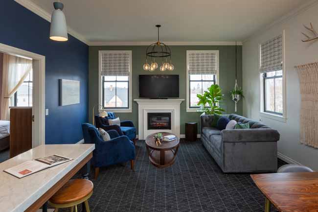 Atticus Hotel Balcony Suite Fireplace Cozy living room with a white fireplace mantel, mounted TV, and two windows letting in light; blue and sage walls with gray sofa and blue chairs.