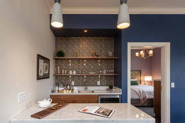 Atticus Hotel Balcony Suite Kitchen Compact kitchenette with a marble countertop, wood drawers, open shelves, and mosaic tile backsplash against a blue wall; doorway to a bedroom on the right.
