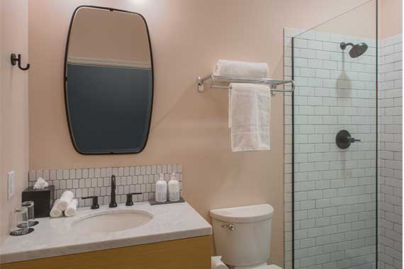 Bathroom with a wooden vanity, white countertop, oval wall mirror, black faucet, towels, toilet, and glass-enclosed shower with white subway tiles.