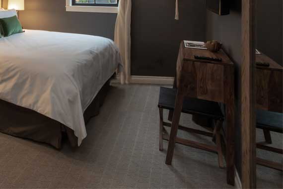 Hotel bedroom corner with a white- duvet bed, dark walls, and a wooden desk with a stool nearby. A window sits above the bed.
