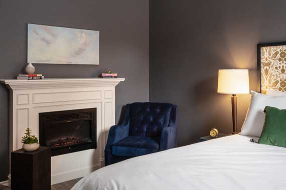 Gray-walled bedroom with a white fireplace, a small plant on a side table, a blue tufted chair, and a bed with a green pillow beside a lit bedside lamp.