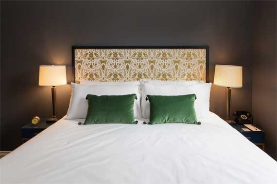 King bed in a dark-toned hotel room with a patterned upholstered headboard, white linens, and two green accent pillows, flanked by identical nightstands and lamps.