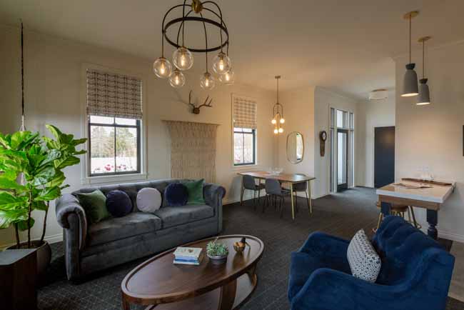 Atticus Hotle Balcony Suite Living Areas Bright open living room with a gray sofa, blue armchairs, a round wooden coffee table, a plant, and windows with woven shades; dining area visible in back.