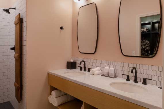 Double-sink bathroom vanity with light peach walls, white hex tile backsplash, black faucets, two oval mirrors, and a wooden open shelf with rolled towels; a walk-in shower with white subway tiles and a wooden towel ladder is visible on the left.