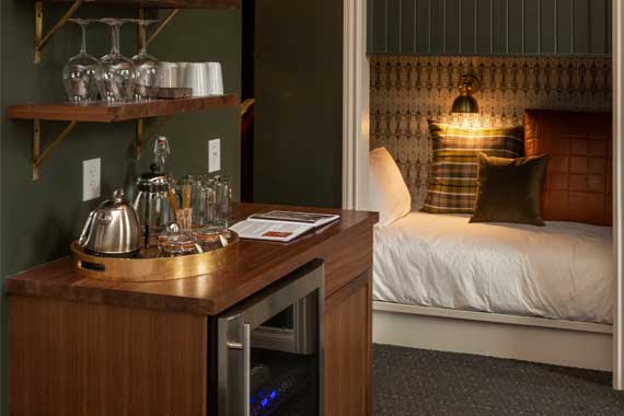 Wooden minibar with a kettle and glassware on top and a microwave beneath; doorway to a bed with plaid and brown cushions in the background.