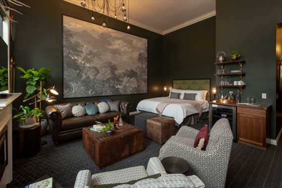 Luxurious hotel suite with dark walls, king bed, brown leather sofa, and a large wall art centerpiece.