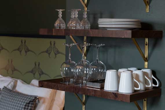 Wall-mounted dark wood shelves with wine glasses, mugs, and plates against a green wall in a cozy hotel room kitchenette.