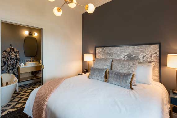 Attiucs-Hotel-Penthouse-Bedroom-to-Bath Hotel bedroom with a king-size bed, gray headboard, and two nightstands flanking lamps; open bathroom area visible to the left.