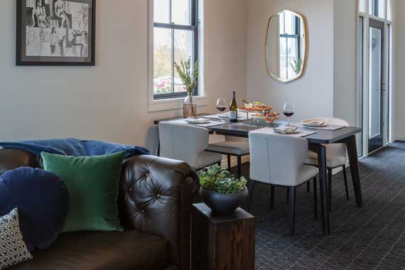 Attiucs-Hotel-Penthouse-Dining-Area Cozy living and dining area with a tufted brown leather sofa, colorful cushions, and a dining table set near the window.