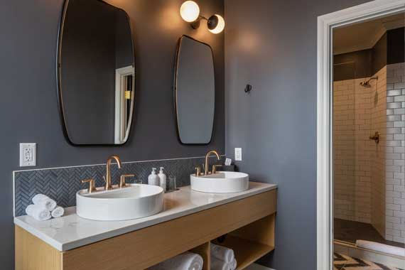 Attiucs-Hotel-Penthouse-Double-Vanity Double vanity with two white vessel sinks and gold faucets, two oval mirrors, gray walls, and a tiled backsplash; shelves with towels and a glass-tiled shower entrance to the right.