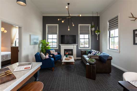Modern living room with blue and brown sofas around a wooden coffee table, a fireplace with a mounted TV on a dark accent wall, and large windows with blinds.