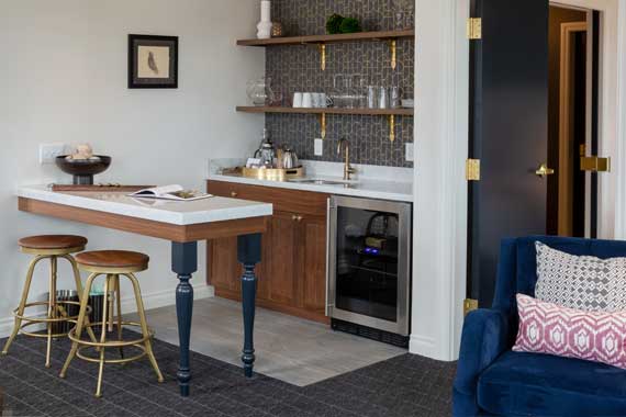 Attiucs-Hotel-Penthouse-Kitchen-Side-view Modern living-room kitchenette with a white island, dark blue legs, two wooden stools, open shelves of glassware, and a stainless steel under-counter cooler; a blue sofa with pink pillows sits nearby.