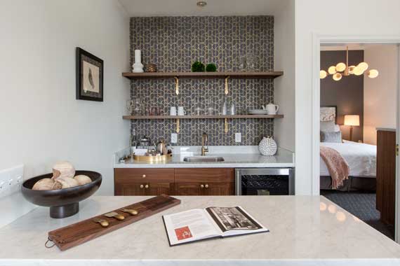 Attiucs-Hotel-Penthouse-Kitchen Compact kitchen area with marble island, wooden shelves, and a tiled backsplash; doorway to a bedroom on the right.