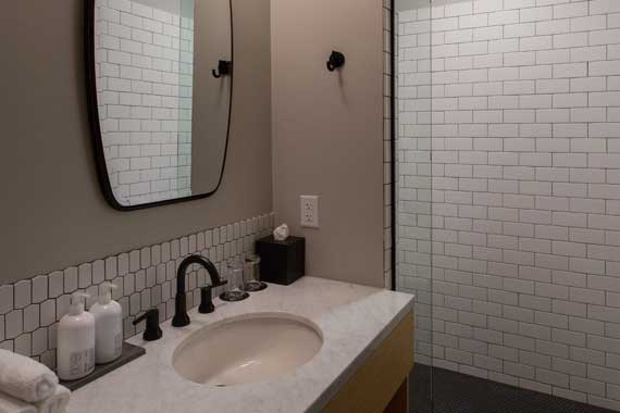 Bathroom vanity area with an undermount sink, black faucet, and light countertop; mirror above and small white subway tiles along the backsplash in a beige-walled bathroom. A tissue box, soap dispensers, and rolled towel sit on the counter, with a glass shower enclosure visible to the right.