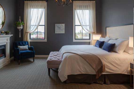 Cozy hotel-style bedroom with dark gray walls, two windows with sheer curtains, a king bed with white linens and blue pillows, and a tufted ottoman at the foot.
