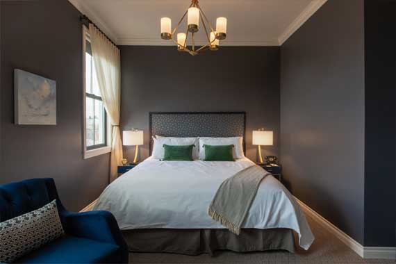 Hotel bedroom with dark gray walls and a white bed featuring a tufted headboard and green pillows, two nightstand lamps, a chandelier, a window with sheer curtains, and a blue armchair.