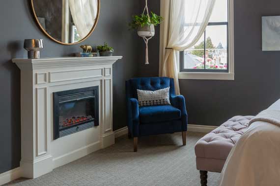 Cozy bedroom corner with a white fireplace, round mirror, blue upholstered chair, hanging plant, and a window with sheer curtains.