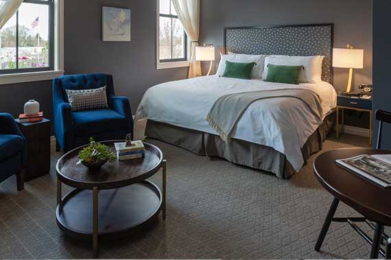 Hotel room with a king bed, two blue armchairs, and a round coffee table by the window. Green throw pillows add color in a modern suite.