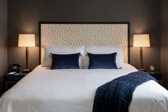 Hotel-style bedroom: white bed with a polka-dotted headboard, two navy pillows, blue knit throw, and symmetrical nightstands with lamps on each side.