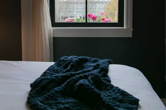 Cozy bedroom scene: white bed sheets with a dark blue knitted throw, window with pink flowers outside and soft natural light.
