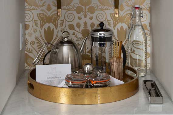 Mini bar setup on a round gold tray with a kettle, French press, water bottle, two glasses, and sugar jars.
