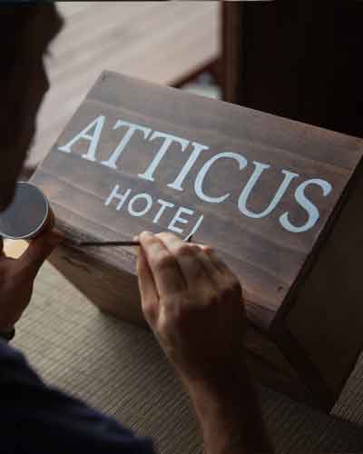 Close-up of a person painting a wooden hotel sign reading 'ATTICUS HOTEL' with a small paintbrush and can nearby.