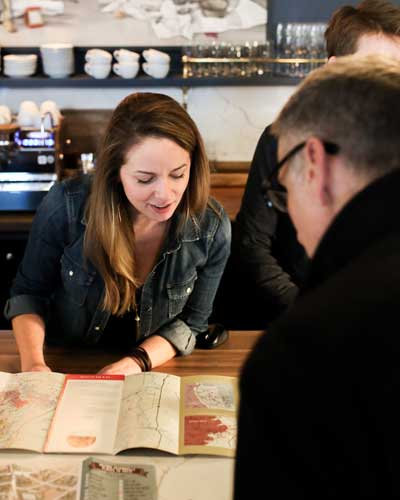 Woman in a denim jacket leans over a map spread on the counter, explaining it to a customer in a cafe setting.