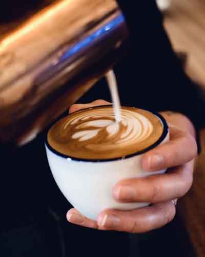 Barista pours steamed milk into a white cup to form latte art, held in both hands with a metal pitcher overhead.