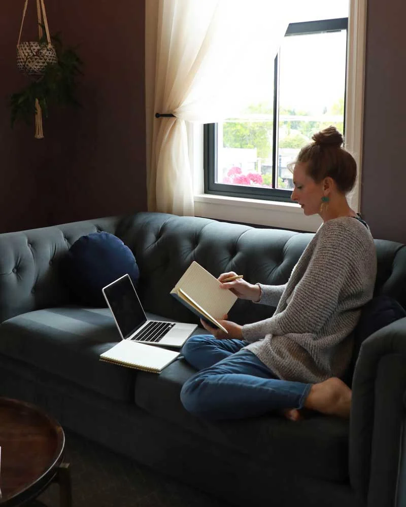 Woman sits cross-legged on a teal tufted sofa, reading a notebook with a laptop nearby by a sunlit window.