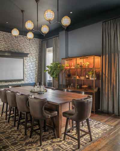 Modern meeting room with a long wooden table, upholstered chairs, hanging pendant lights, a patterned rug, and a wooden bookshelf along dark walls.