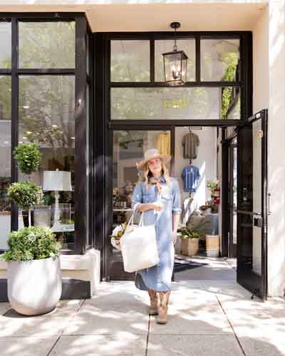 Woman in a denim dress and hat walking out of a boutique with a white tote bag and ankle boots, doorway framed by black glass panels outside a storefront.