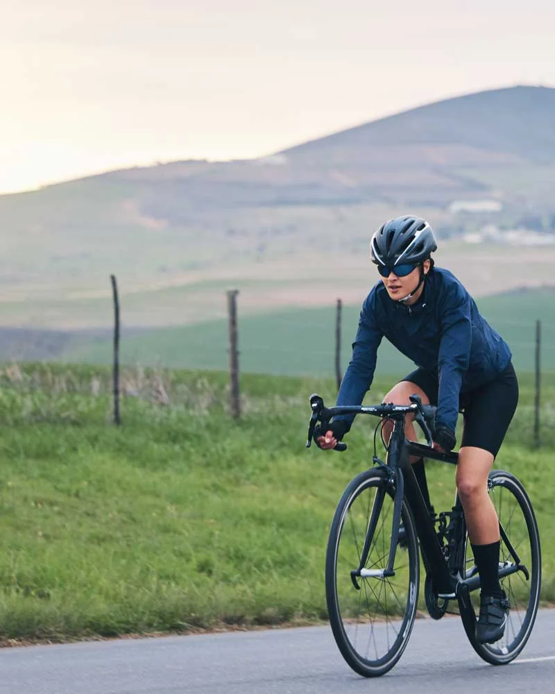 Cyclist wearing a helmet and sunglasses rides a black road bike along a rural country road with hills in the background.