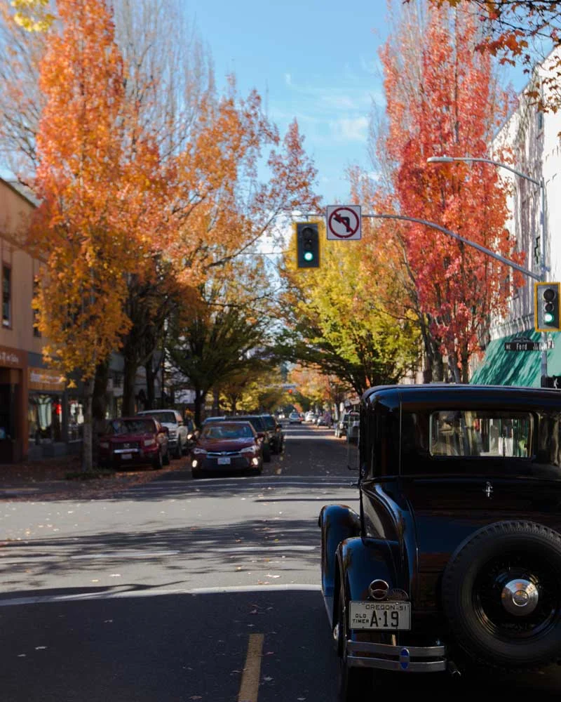 Autumn city street with orange and yellow trees, a vintage black car parked at the curb on the right, and a green traffic light overhead.