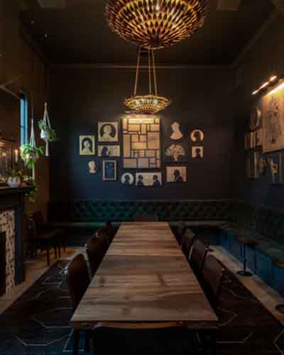 Dimly lit dining room with a long wooden table, black chairs, a teal banquette along the back wall, and a circular chandelier overhead.