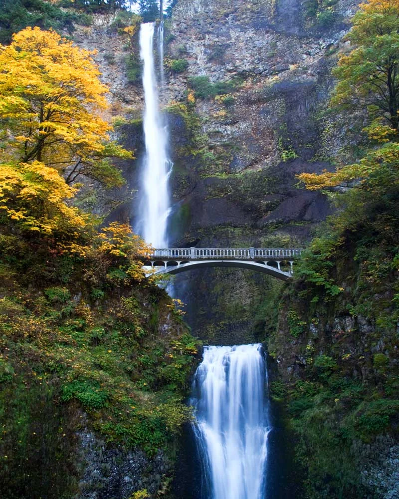 Tall waterfall cascading down a rocky cliff with a stone arched bridge, autumn trees in yellows and greens.