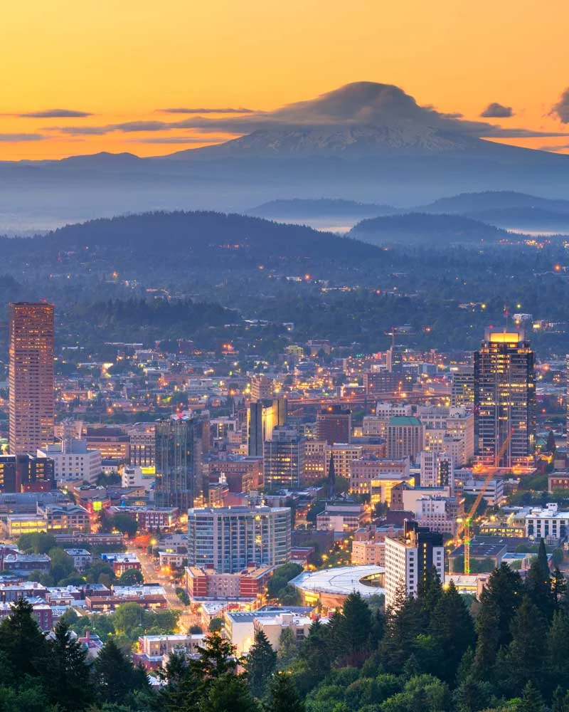 City skyline at sunset with a distant mountain range and layered hills fading into a hazy blue horizon.
