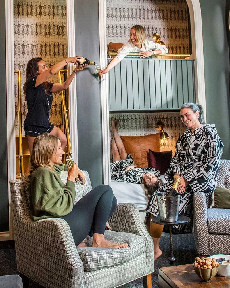 Group of friends in a stylish living room celebrate with champagne, one woman on a balcony pours to others below.