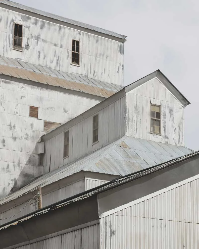 Atticus-Hotel-Granary-District Weathered white metal warehouse with multiple gable roofs and small rectangular windows, showing peeling paint.
