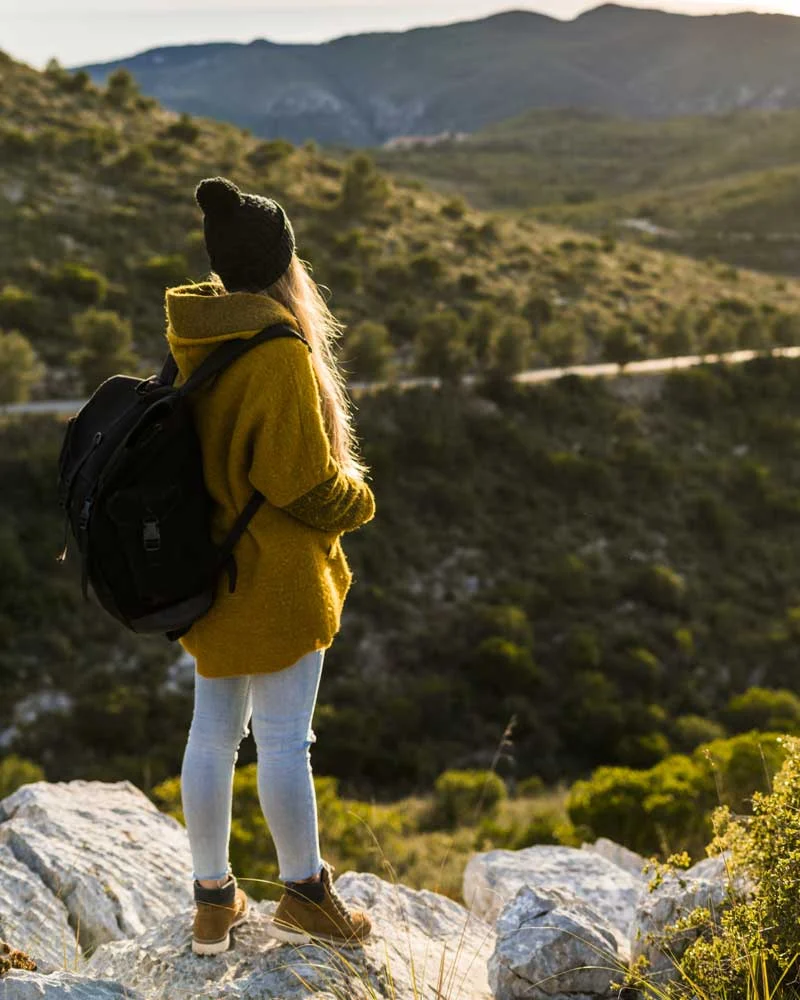 Hiker in a yellow sweater and beanie stands on rocky overlook with a backpack, gazing at a mountainous landscape.