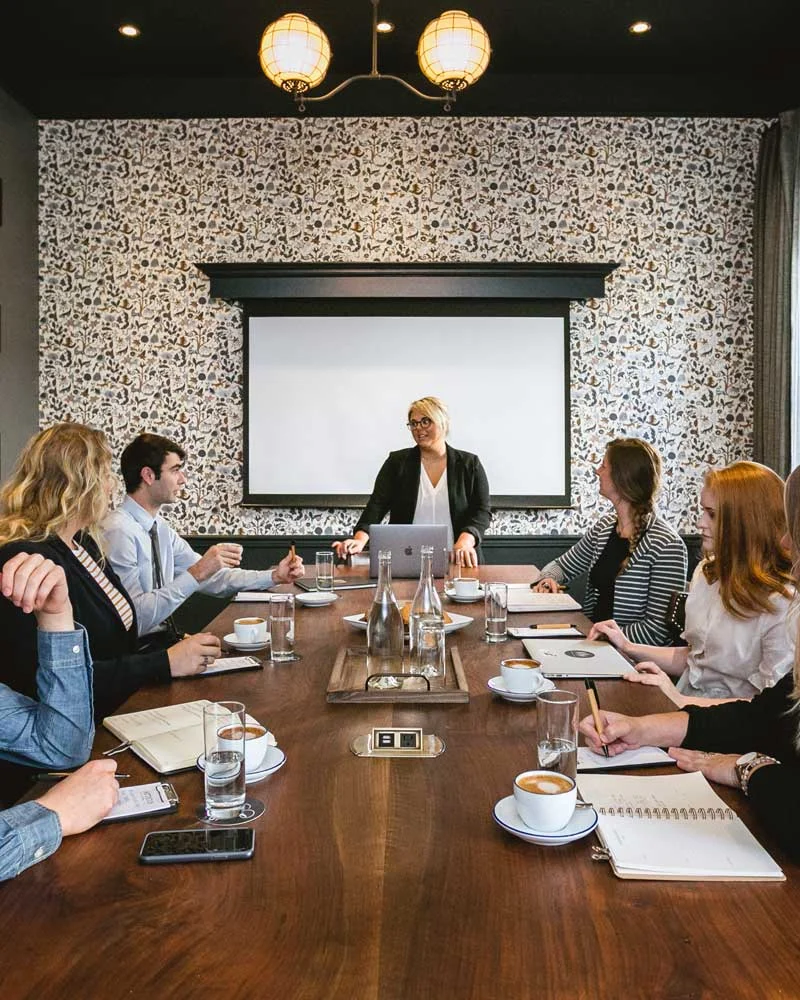 Professional meeting around a long wooden table as a woman presenter stands by a projector screen at the front.