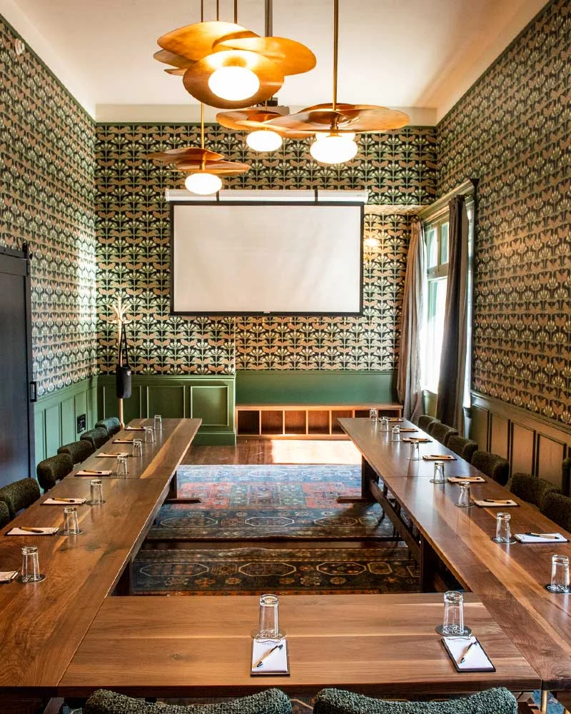 A formal meeting room with long wooden tables arranged in a U-shape, green wainscoting, ornate wallpaper, and a projector screen at the front.