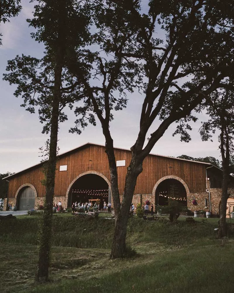 Rustic wooden barn with arched entryways and string lights, guests gathered outside among trees at dusk.