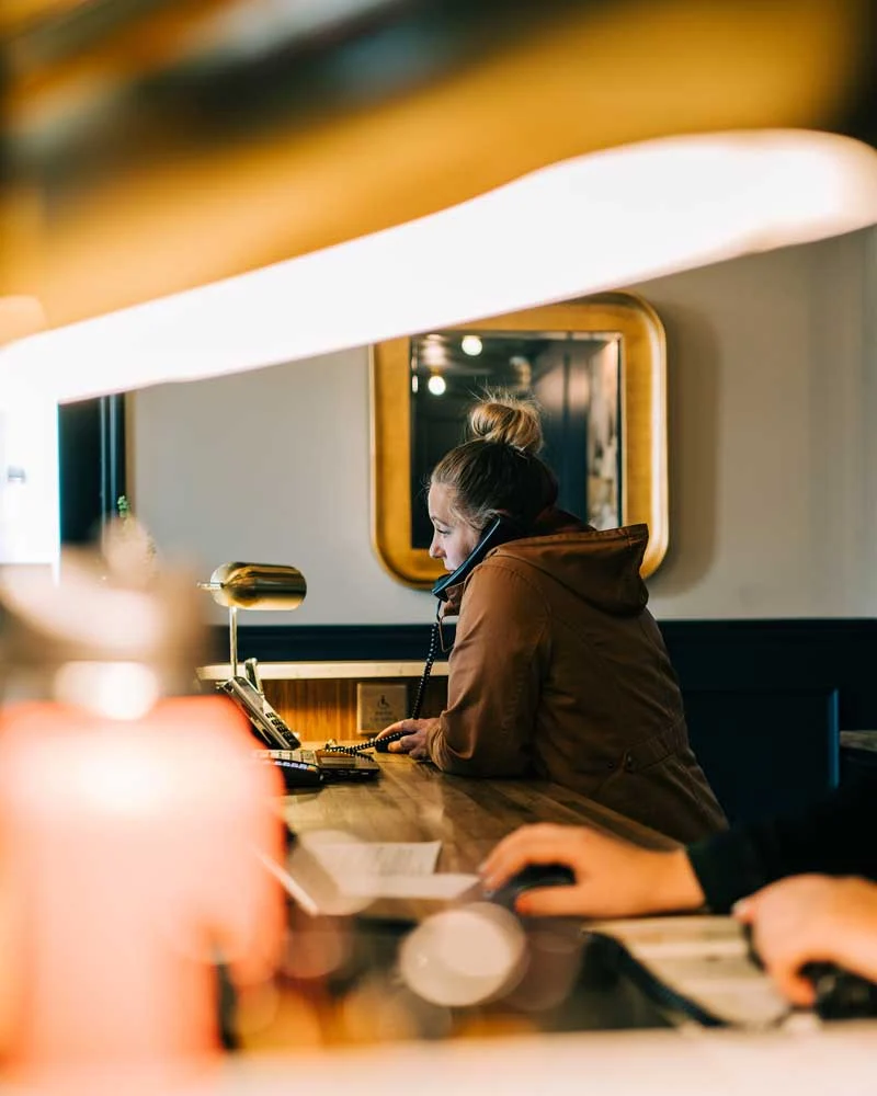 Receptionist wearing a brown jacket with a headset, seated at a wooden desk and using a phone.